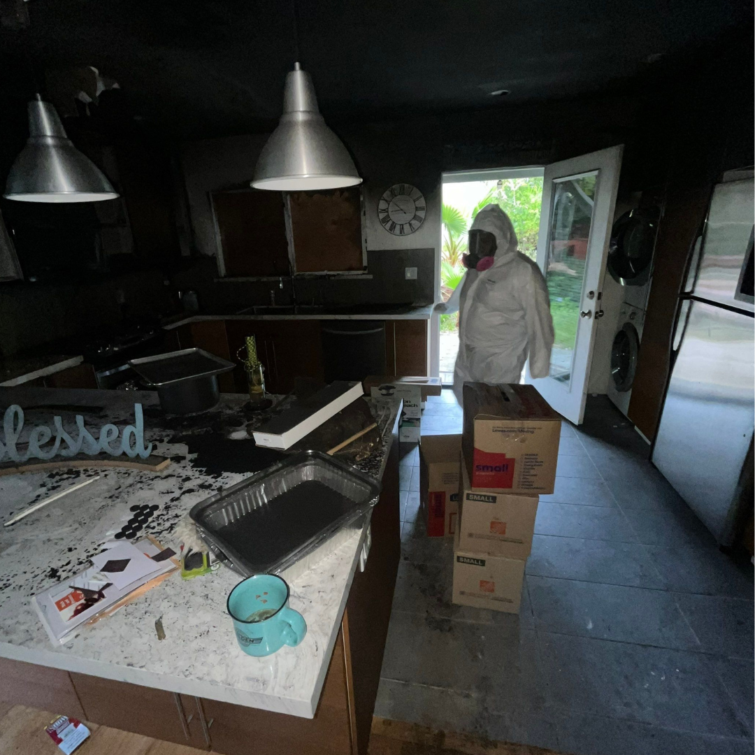 A worker in protective clothing inside a soot-damaged kitchen.