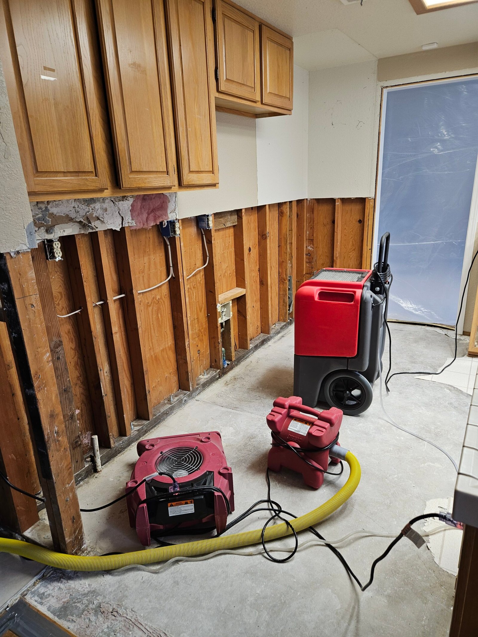 Drying equipment beside exposed kitchen wall studs and a plastic containment barrier.